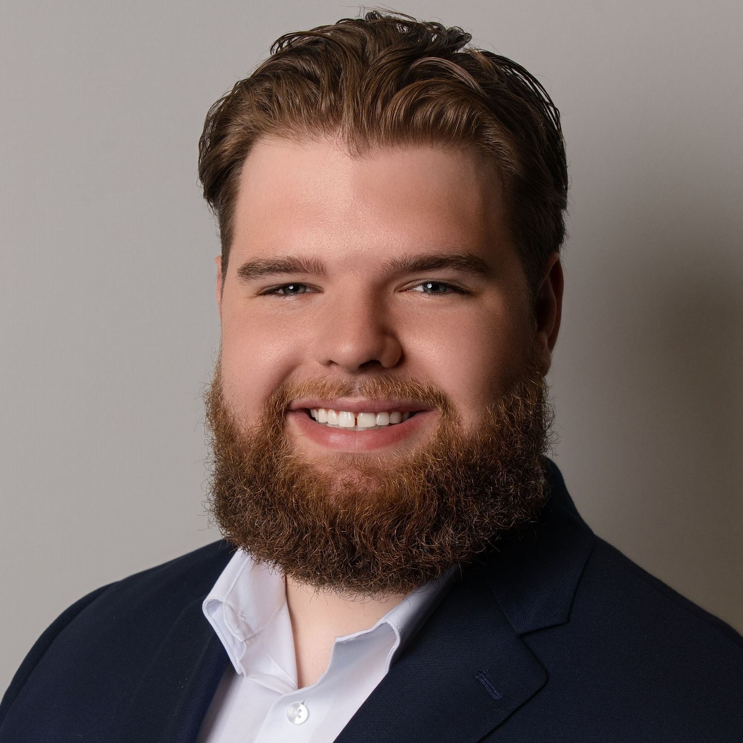 Professional headshot of a person with a full beard wearing a navy suit and white dress shirt against a neutral background.