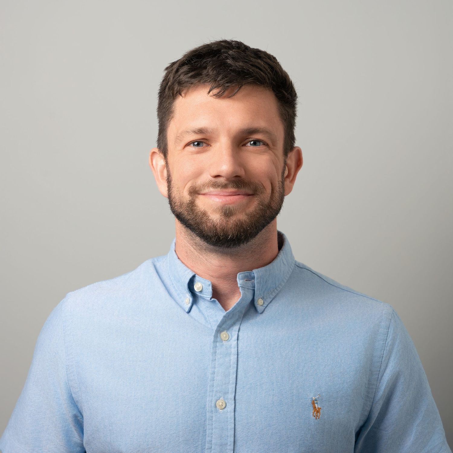 Professional headshot of a person wearing a light blue button-down shirt against a neutral gray background.