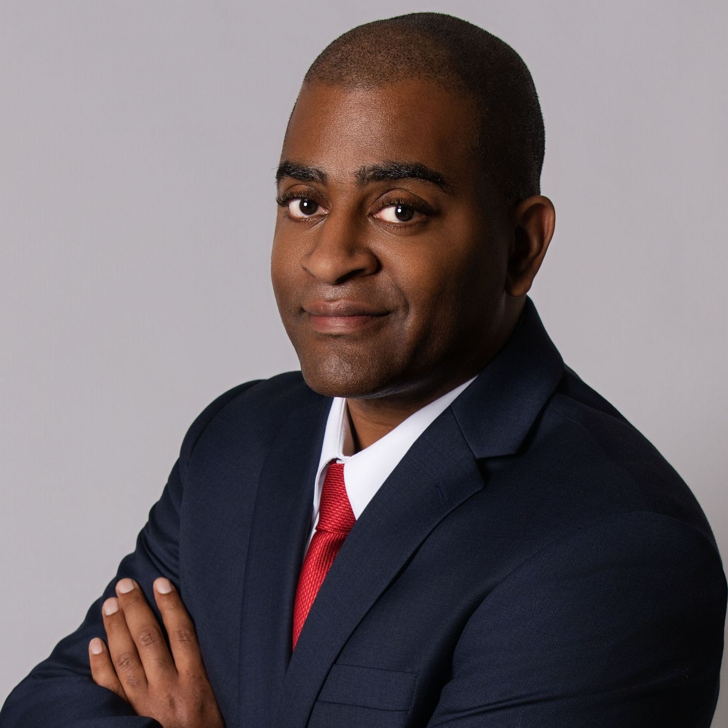 Professional headshot of person in navy suit and red tie with arms crossed against gray background.