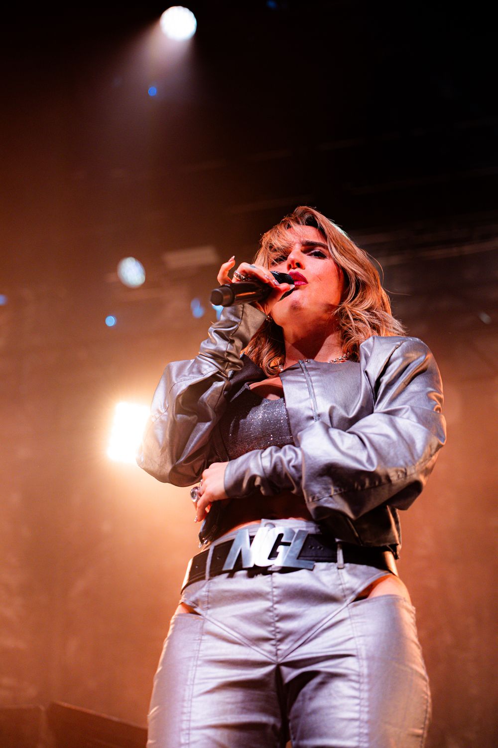 Concert performer in white outfit energetically singing on stage with warm lighting.