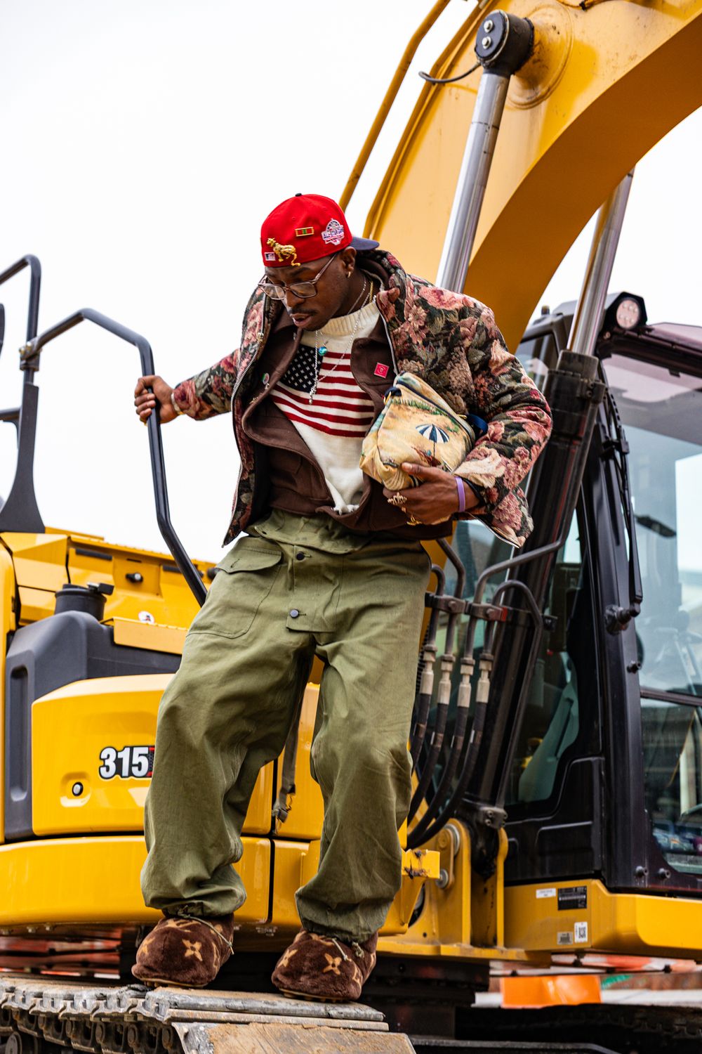 Construction worker in safety gear stands next to yellow excavator equipment on job site.