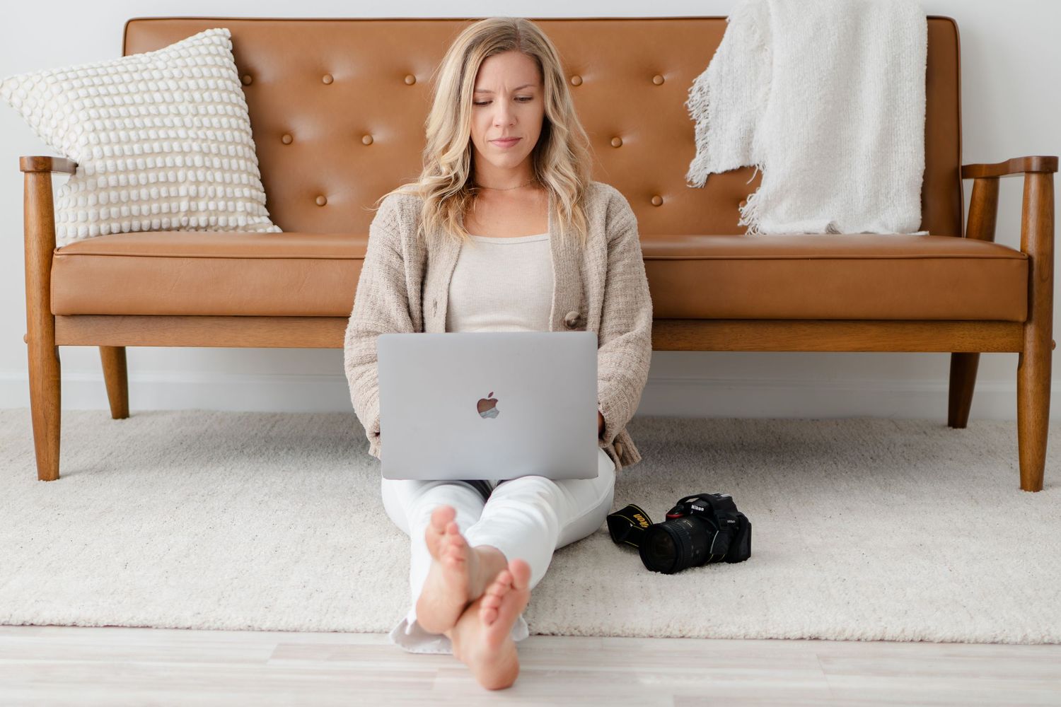 Someone wearing casual clothing works on a MacBook laptop while sitting on the floor against a brown leather sofa.
