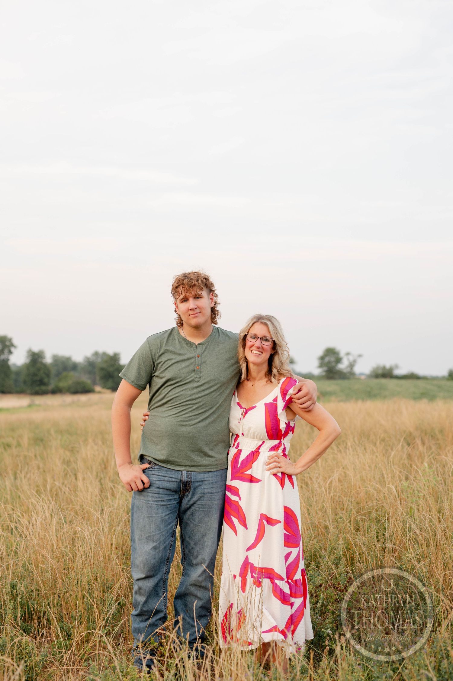 A couple poses together in a golden field at sunset, she wearing a pink floral maxi dress and he in casual green.