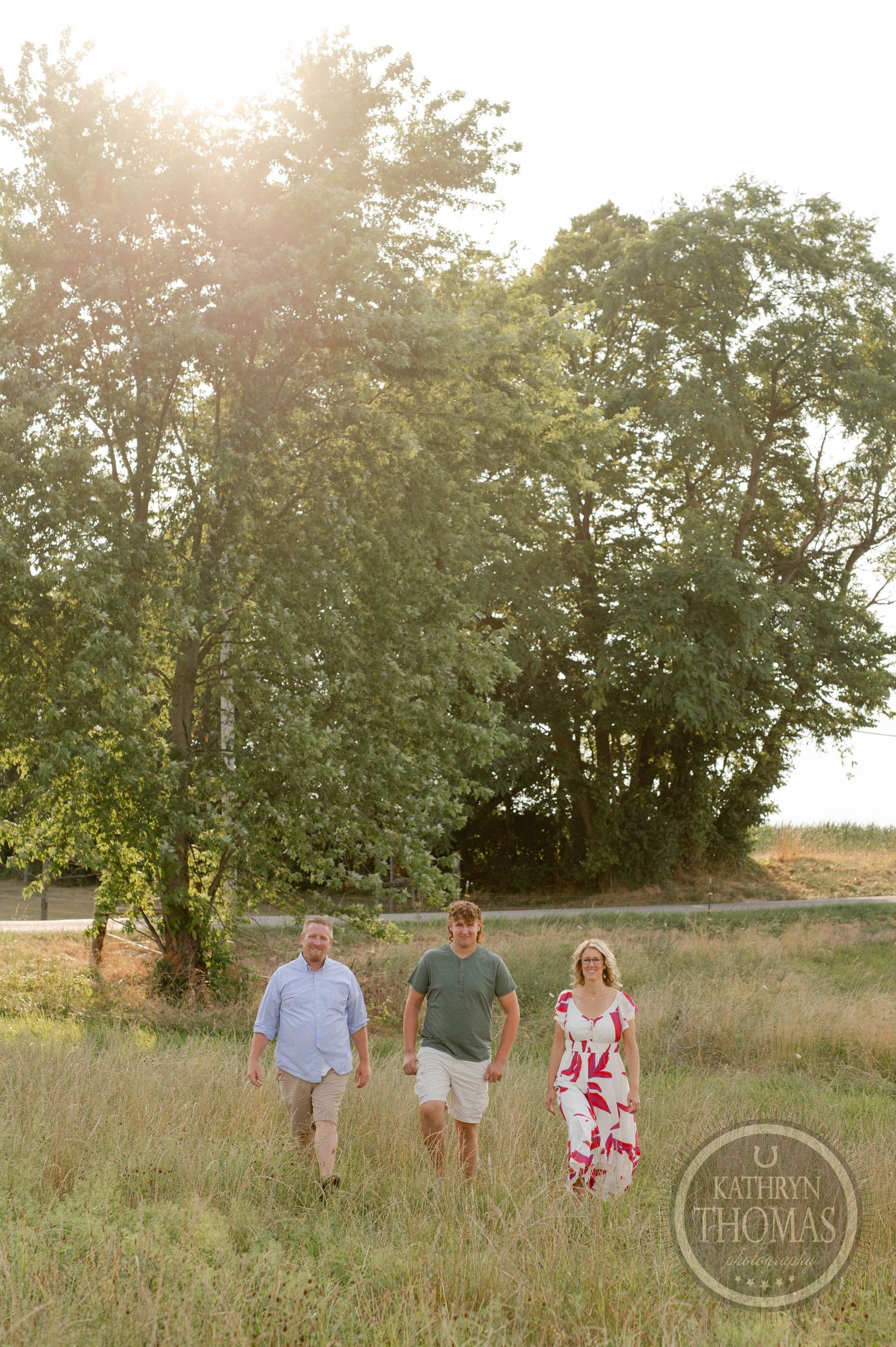 Three people walking through tall grass with sunlit trees in the background.