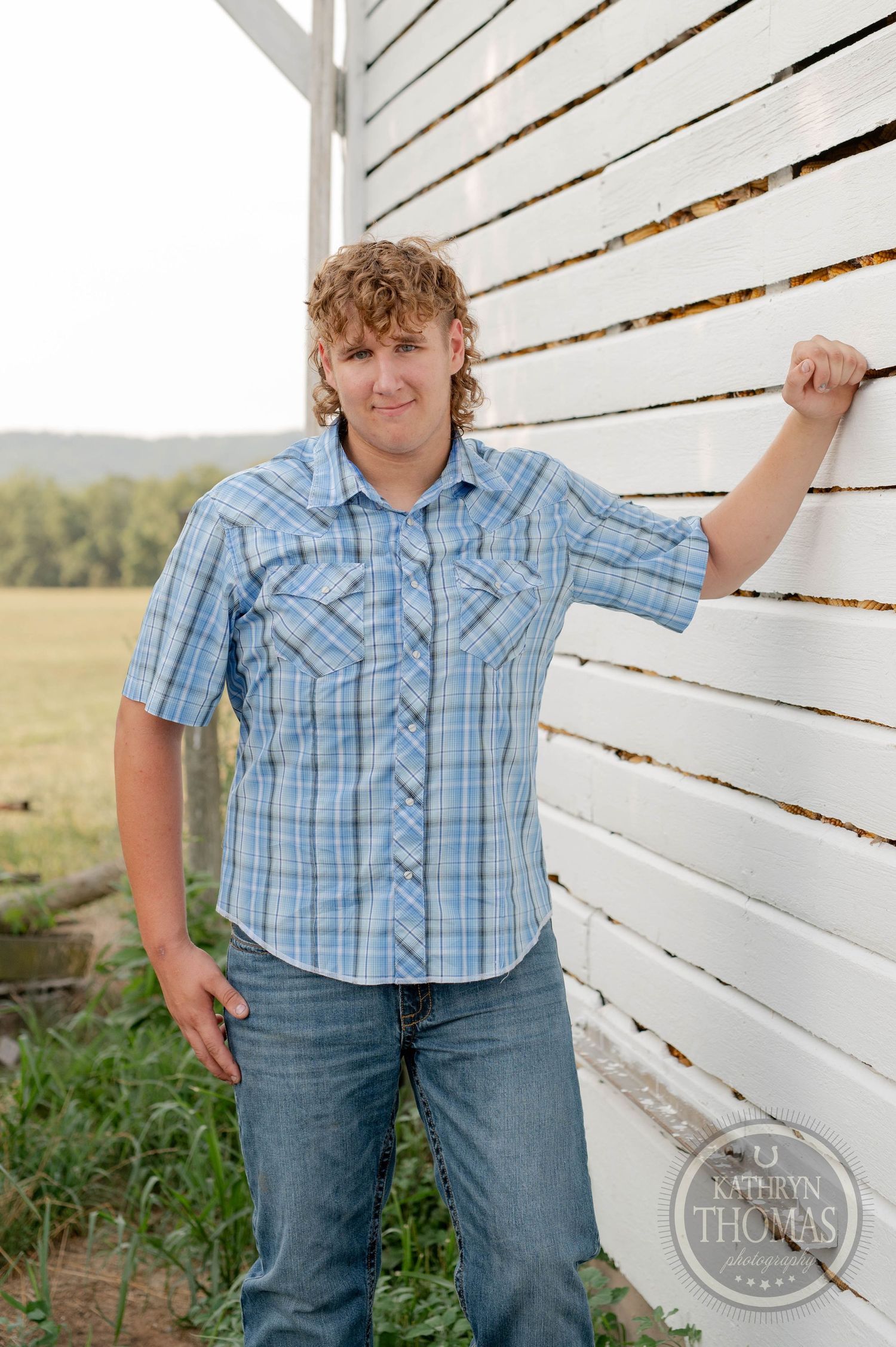 A casual country portrait against a white wooden barn wall wearing a blue plaid shirt and jeans.
