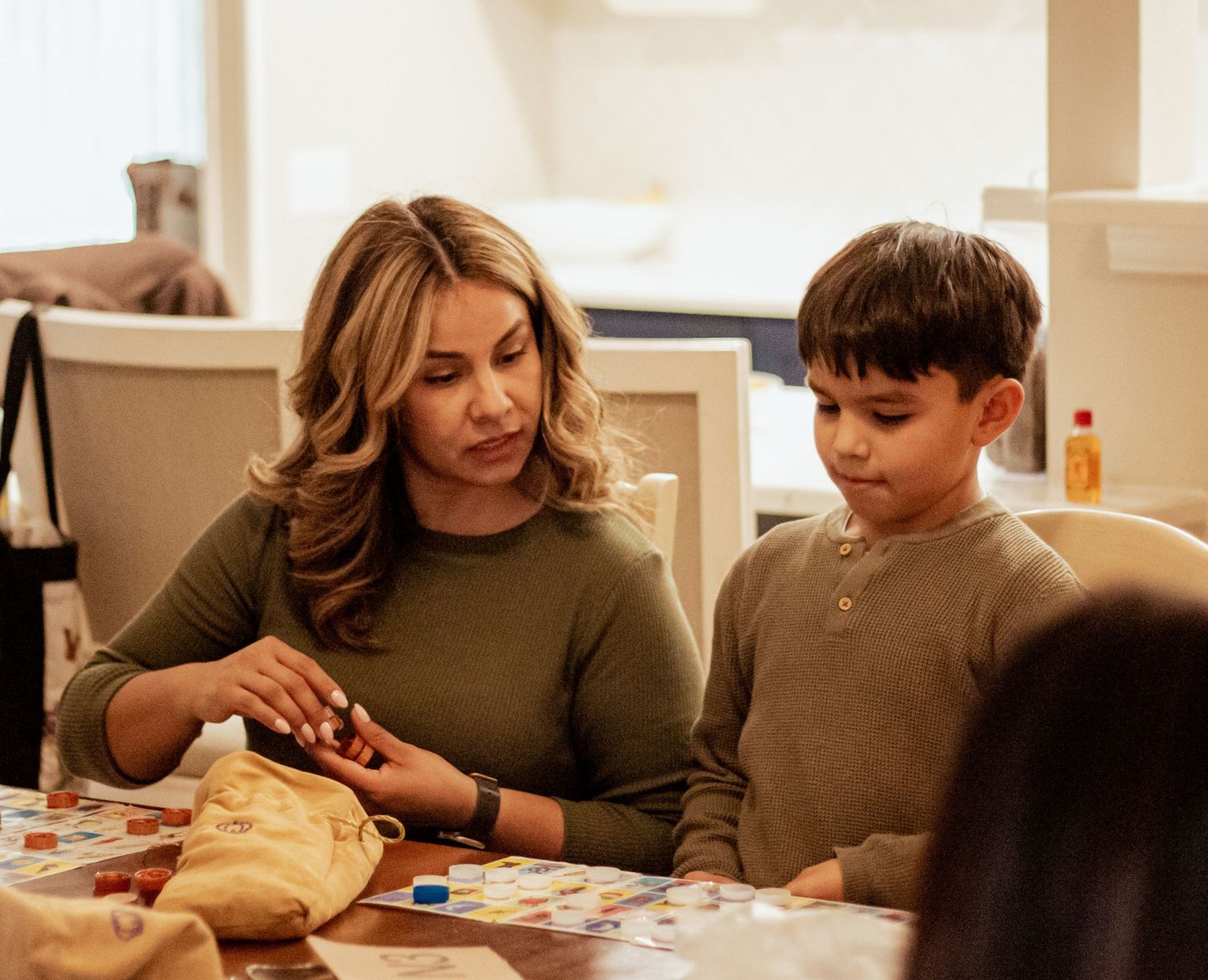 A parent and child sit together at a table working on crafts or activities in a warm-lit home setting.