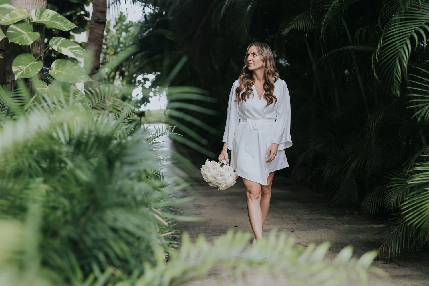Woman in white robe walking through a lush tropical garden path.