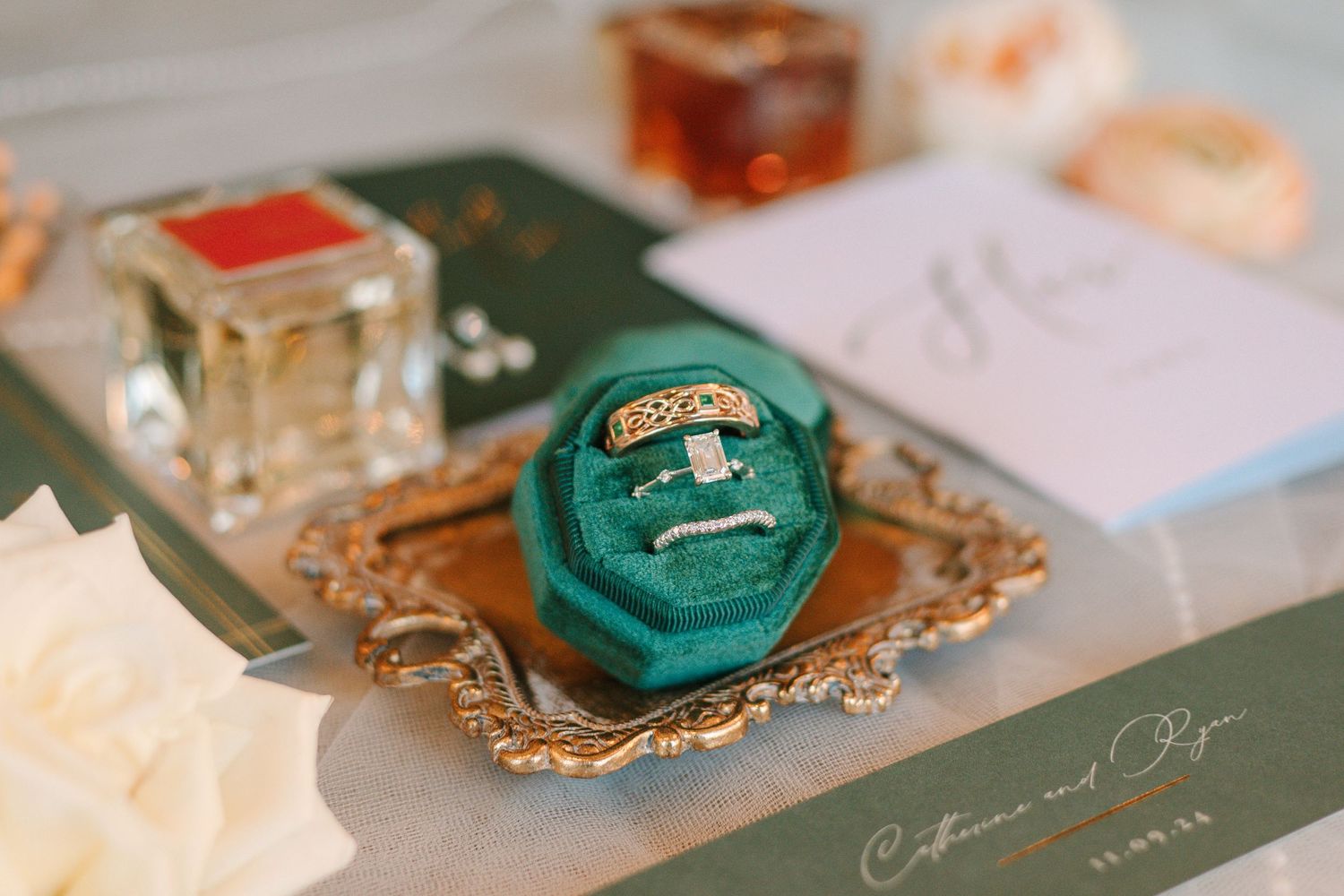 A teal velvet ring box sits on an ornate gold tray with wedding stationery and decor items nearby.