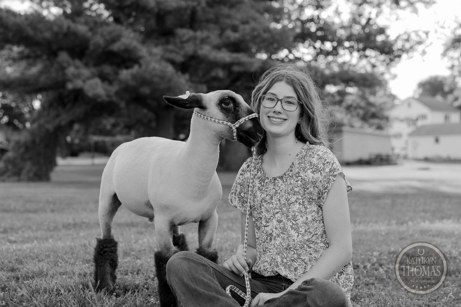 A black and white portrait showing a young person in glasses sitting outdoors with a show lamb on a lead.