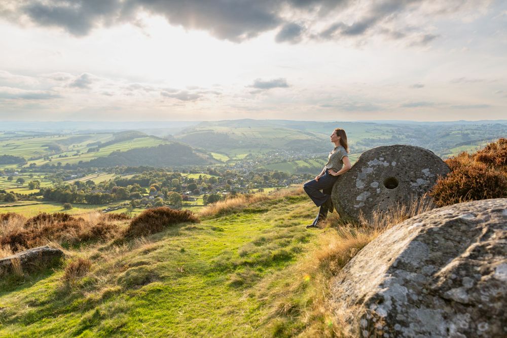 Someone rests on rocky hilltop overlooking scenic green valley with dramatic cloudy sky.