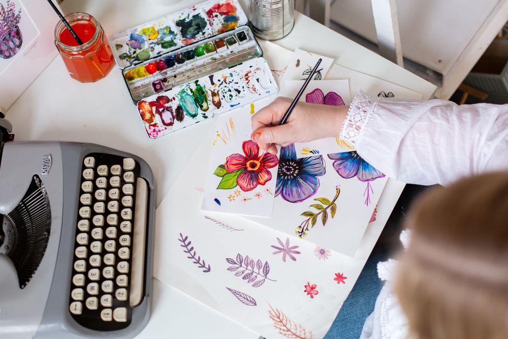 Watercolor painting supplies and vintage typewriter on white desk with colorful artwork.