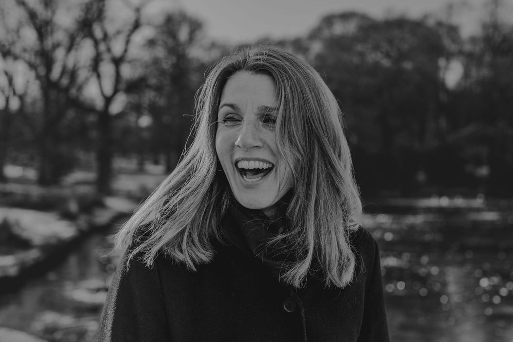Black and white portrait of someone laughing joyfully outdoors with windswept hair and wearing a dark sweater.