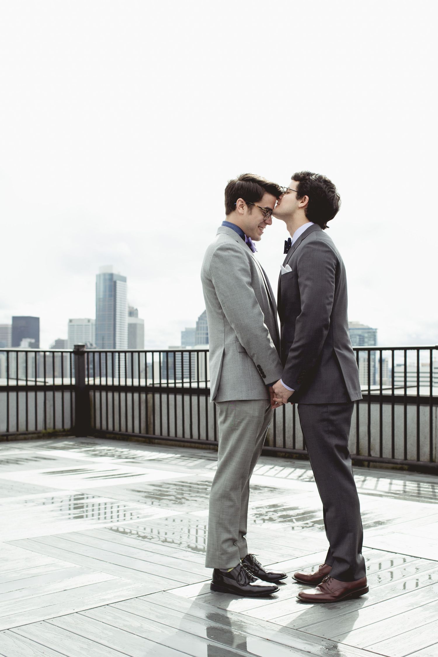 Two people in suits share a romantic moment on a rooftop deck with city buildings visible in the background.