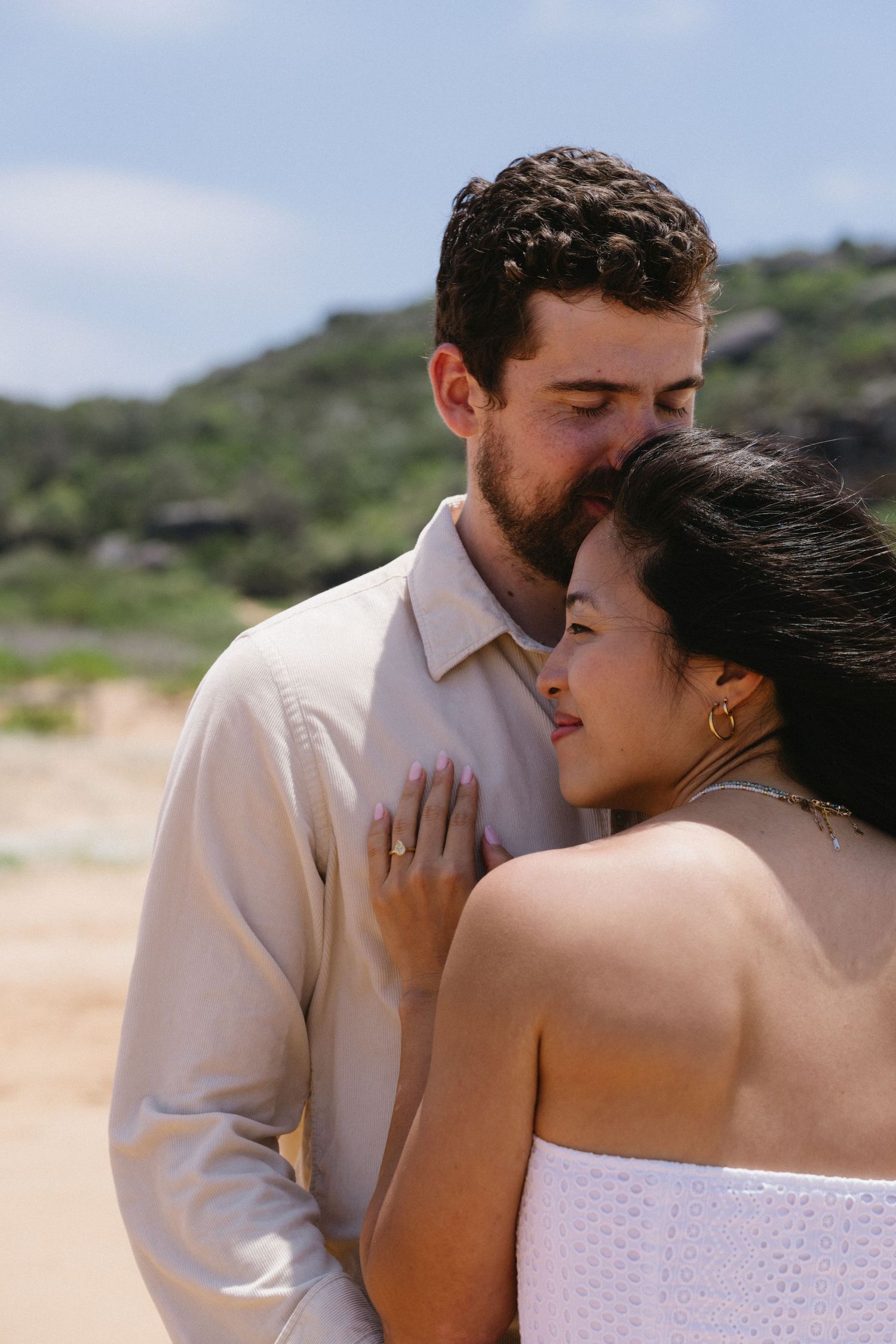 Intimate portrait of couple embracing on sunny beach with natural coastal landscape.