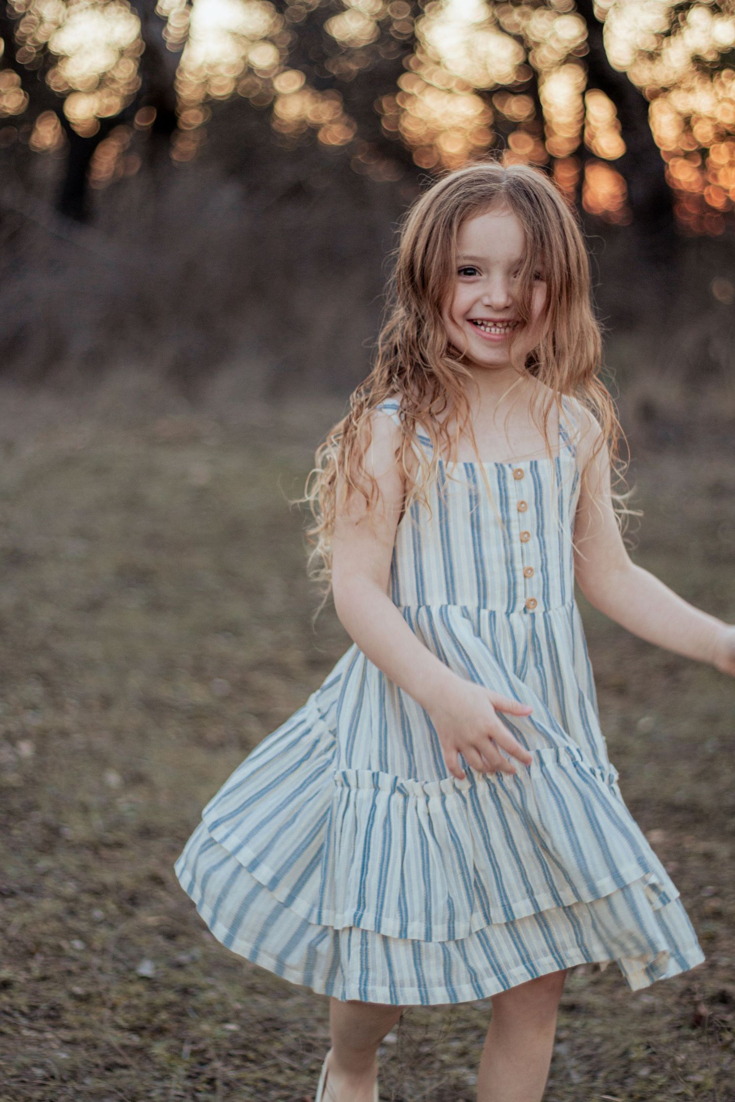 Young child in blue striped summer dress twirling and laughing outdoors at sunset.