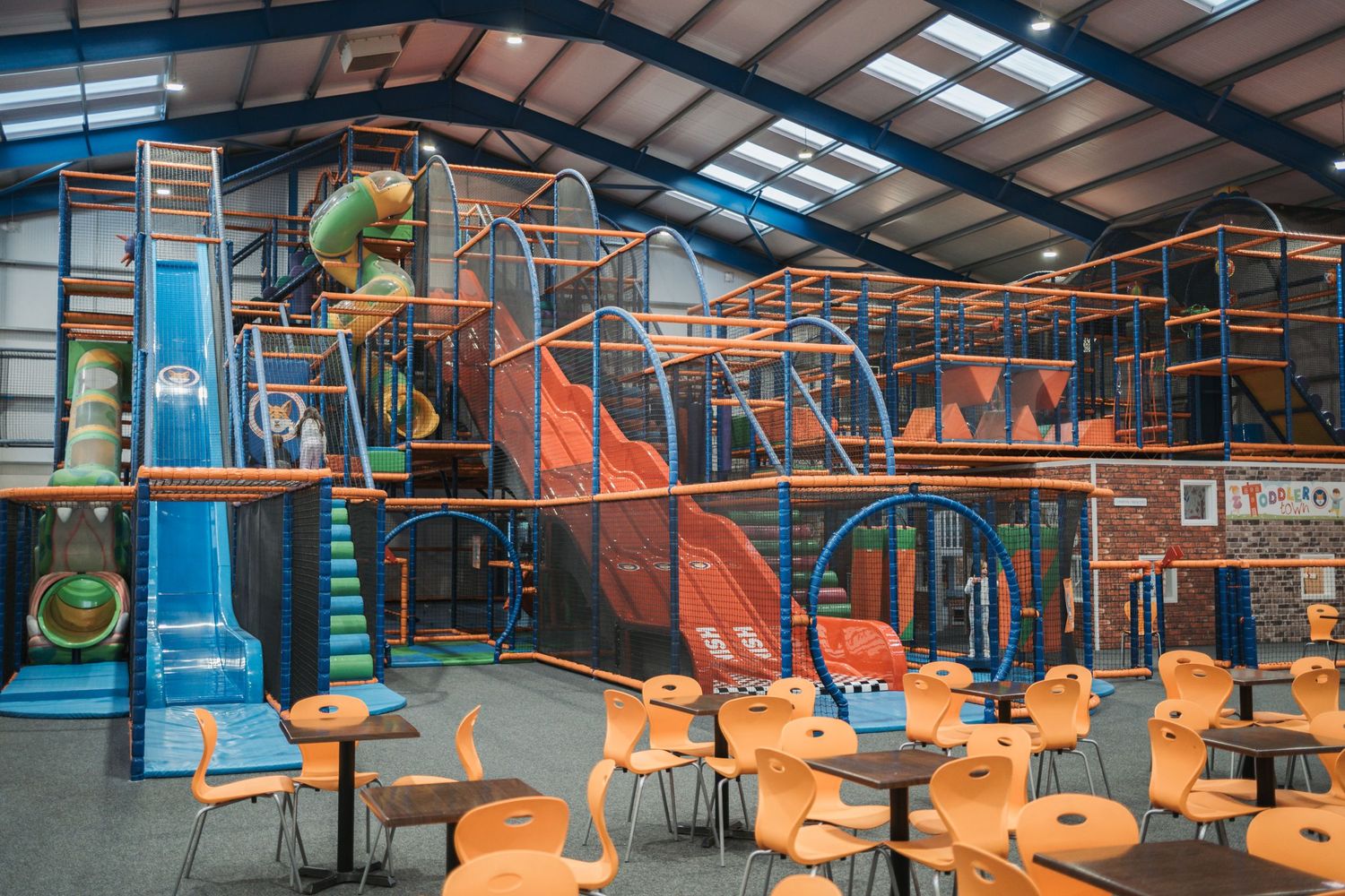 Large indoor playground structure with orange slides, climbing equipment and seating area in a spacious warehouse setting.