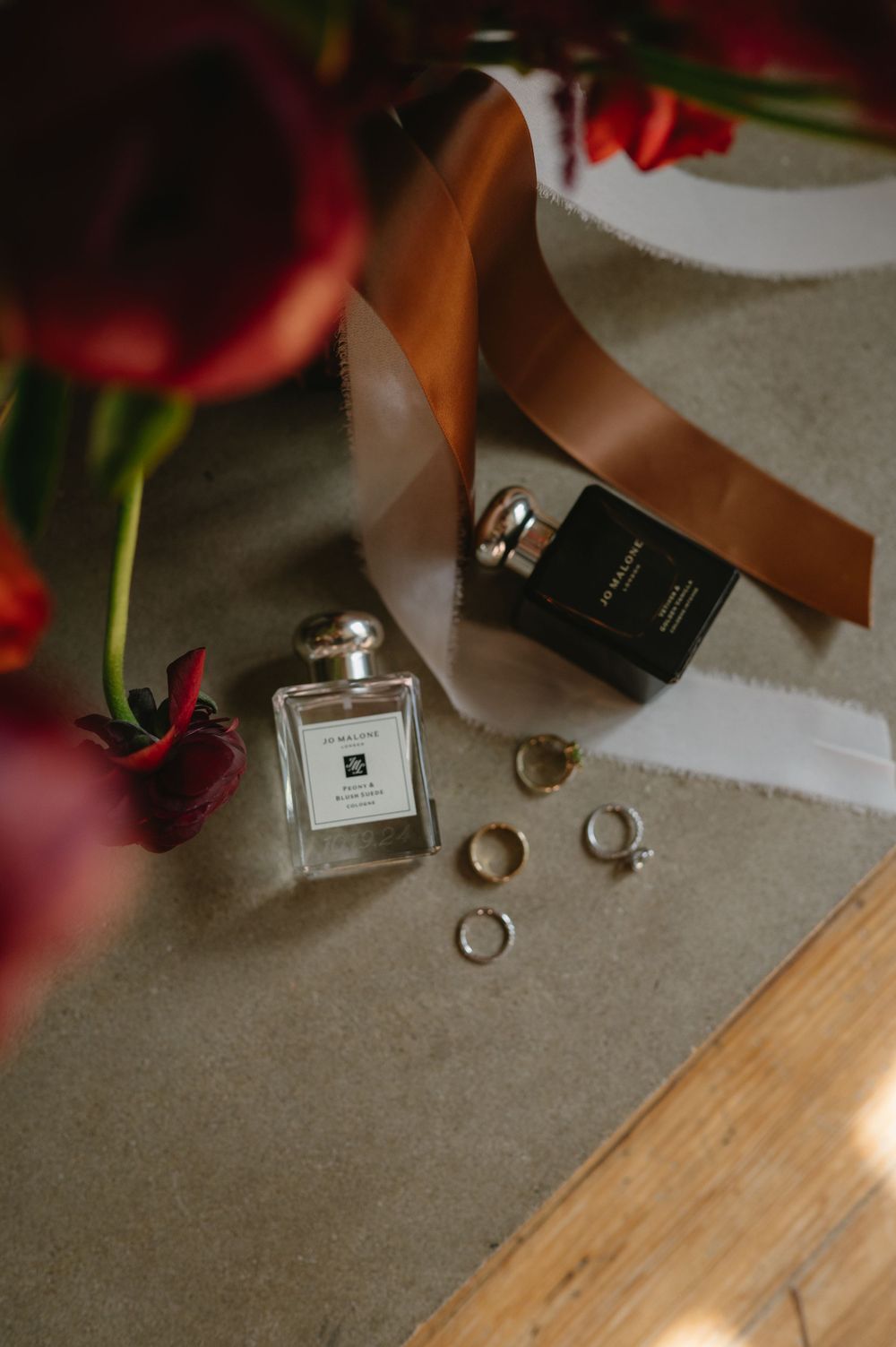 Wedding rings and perfume bottles arranged with red roses on a neutral surface.
