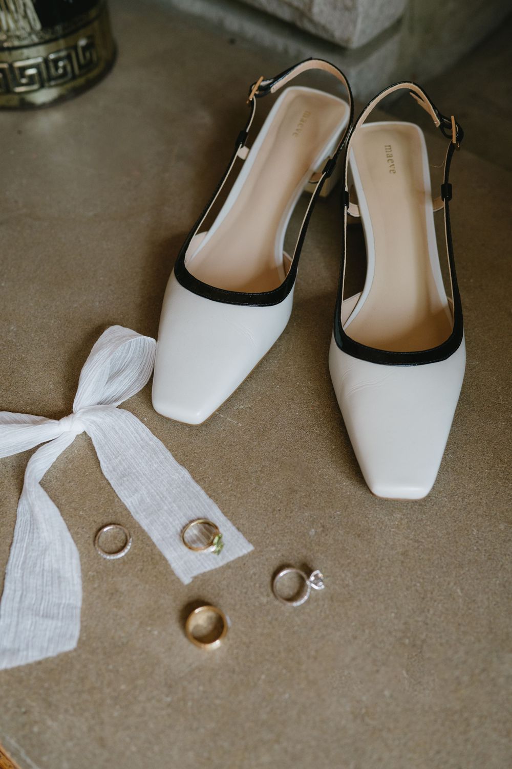 White and black slingback shoes with wedding rings and a white ribbon on a beige surface.
