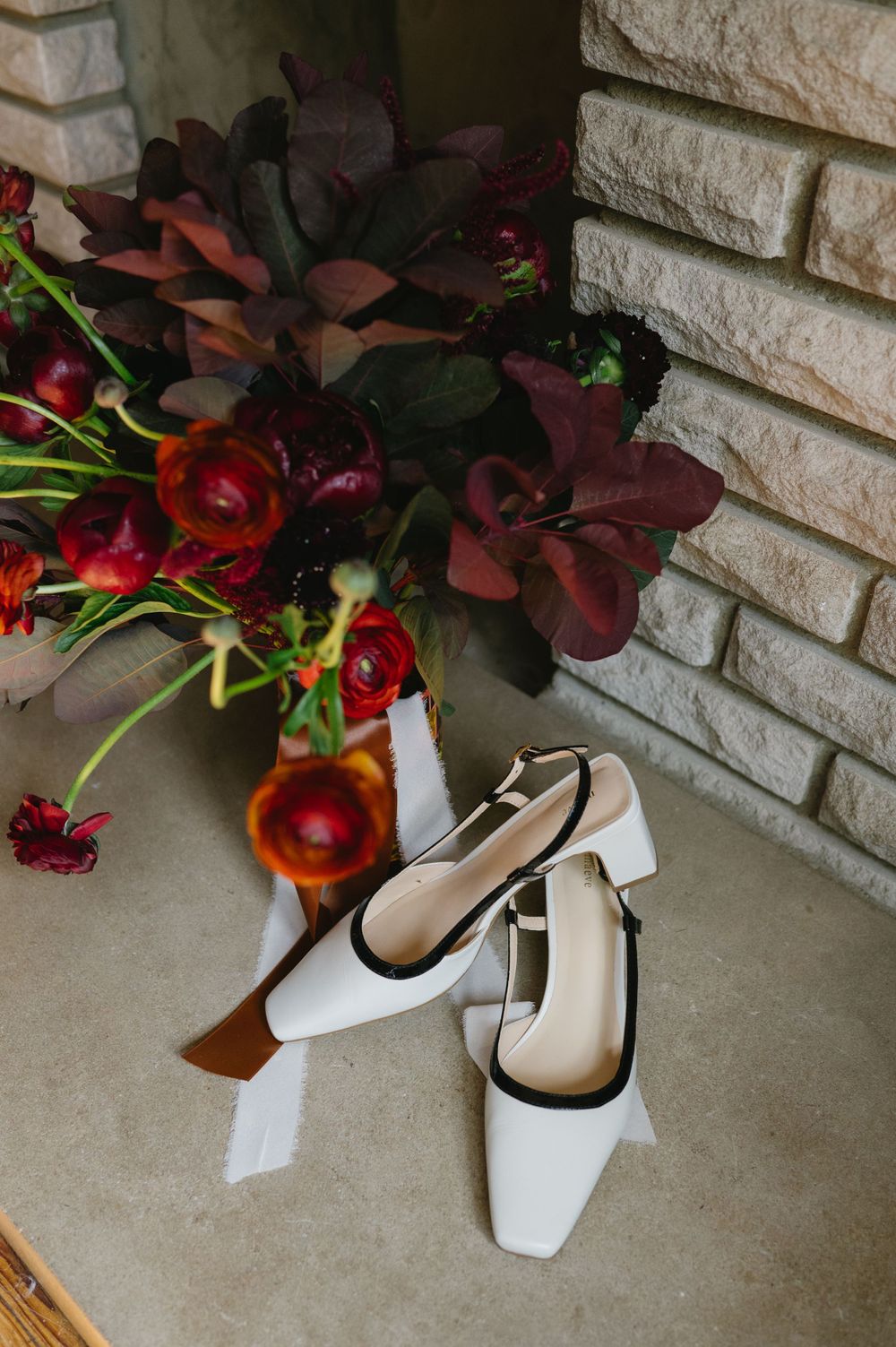 Elegant white bridal shoes placed near a vibrant burgundy floral arrangement against stone wall.