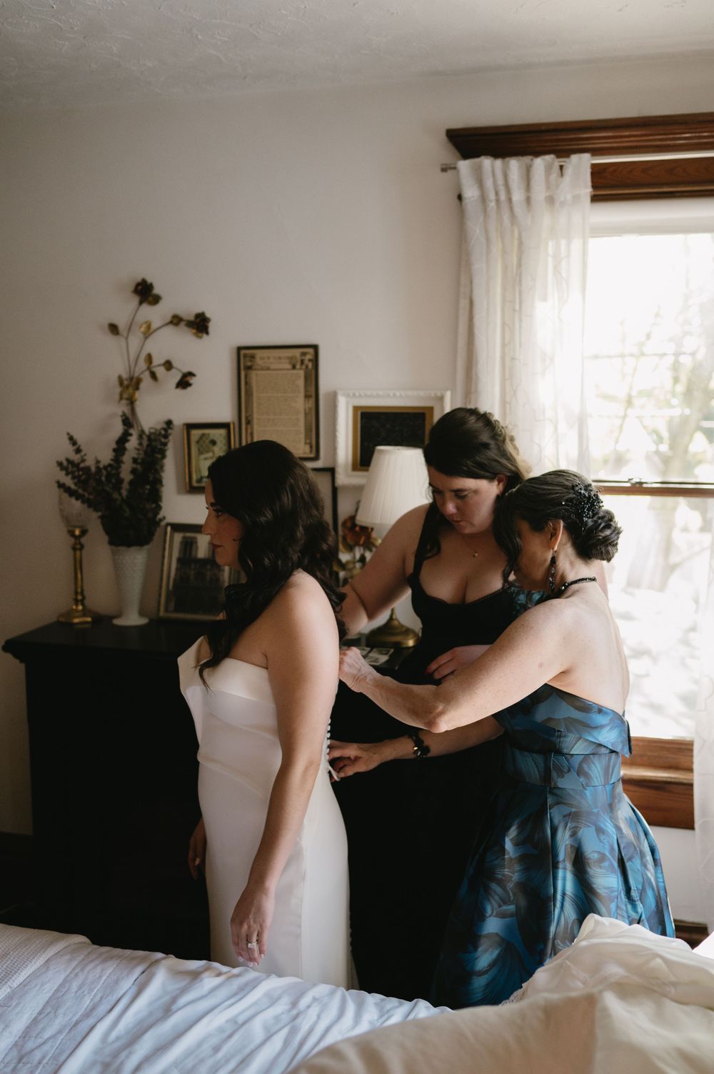 Bridesmaids helping with wedding dress preparations in a sunlit room.