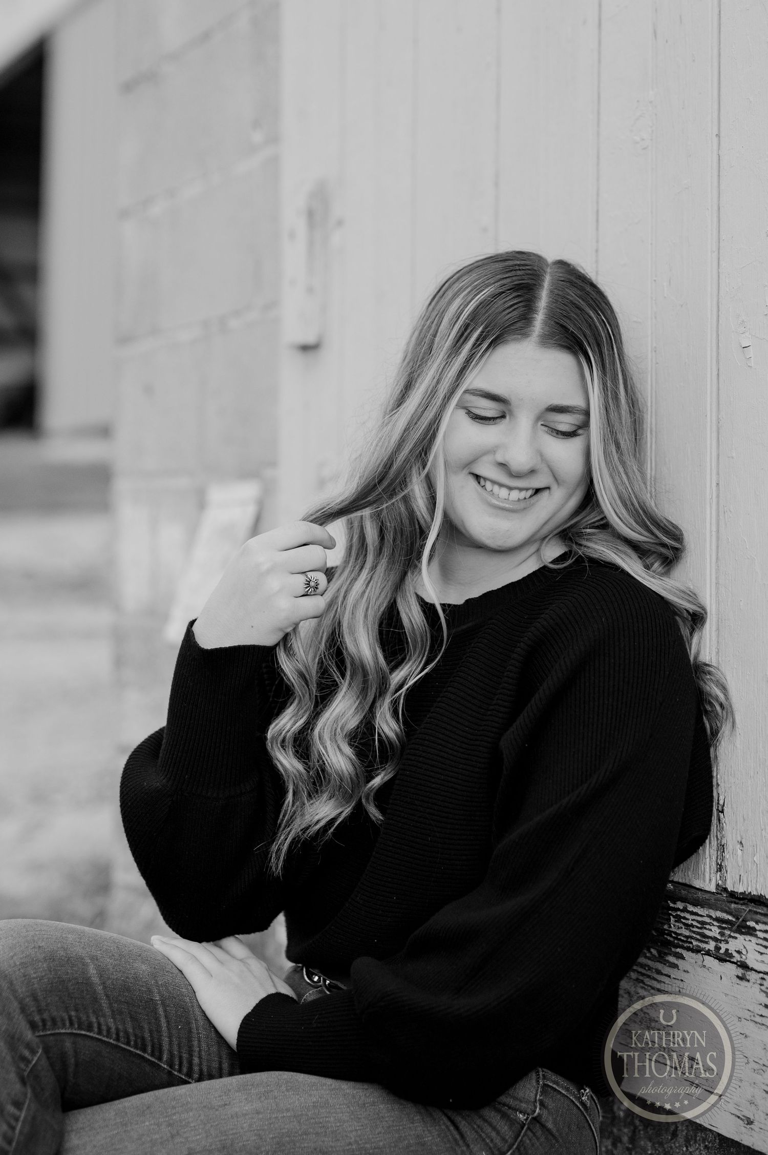 Black and white portrait showing casual style with wavy hair and cozy sweater against rustic wall.