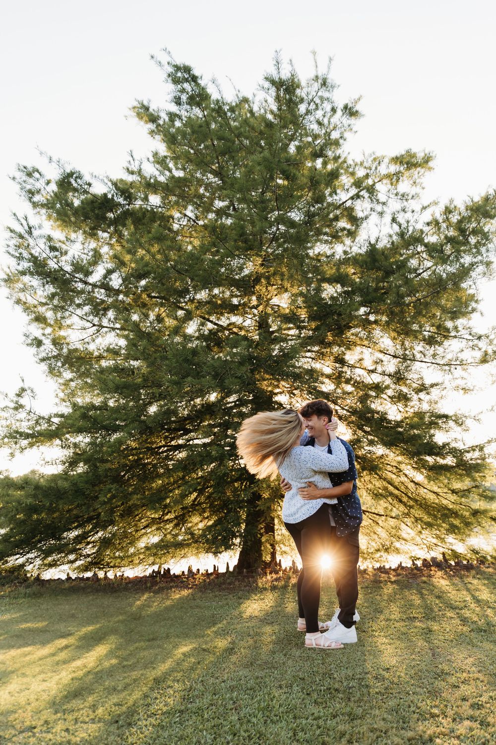 A couple embraces in front of a large tree during golden hour with sunlight streaming through the branches.