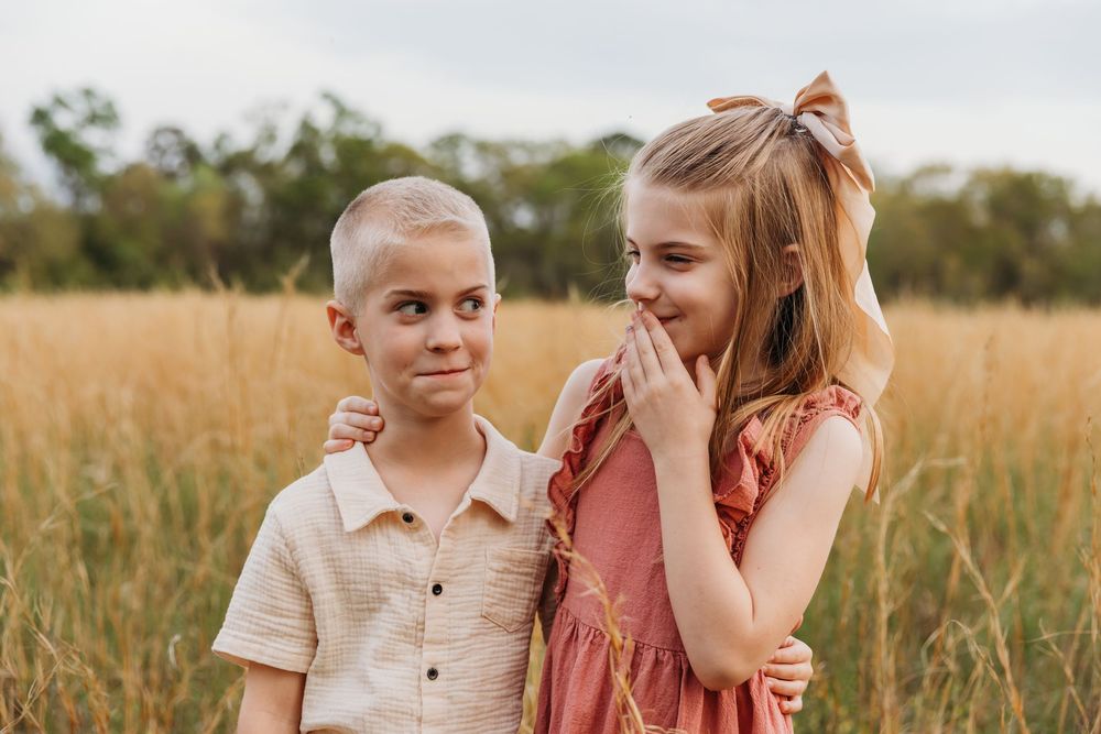 Two young children pose together smiling in a sunlit golden field during a summer evening.