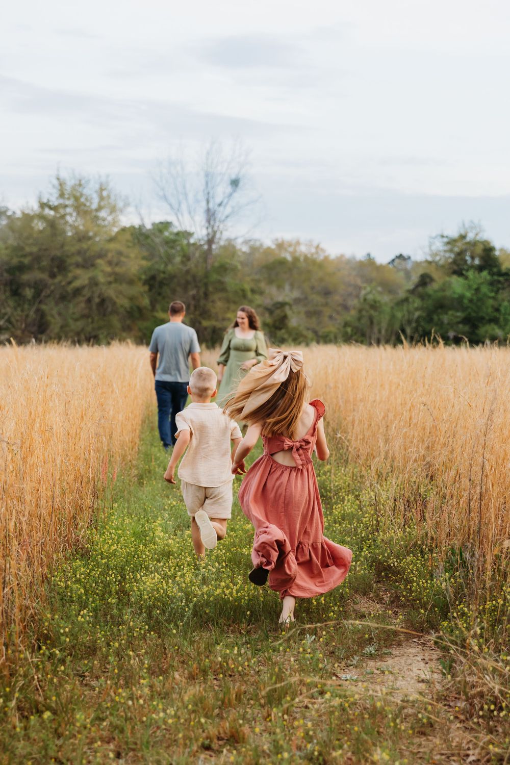People walking through a golden wheat field on a grassy path surrounded by autumn foliage and trees.
