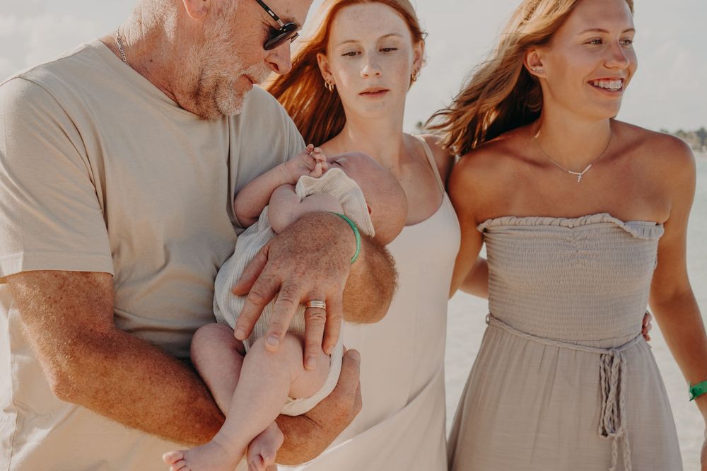 Family at beach with grandfather holding newborn baby, red-haired mother and teenage daughter smiling nearby.