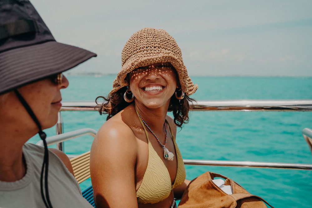 Two women wearing hats smile on a boat with turquoise ocean water in the background.