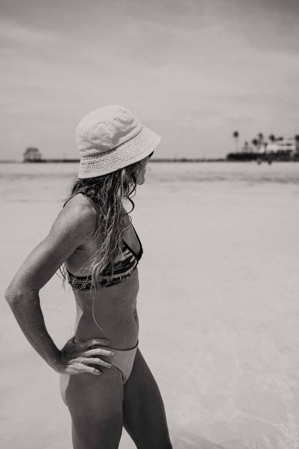 Black and white photo of a woman in a bikini and bucket hat standing on a tropical beach.