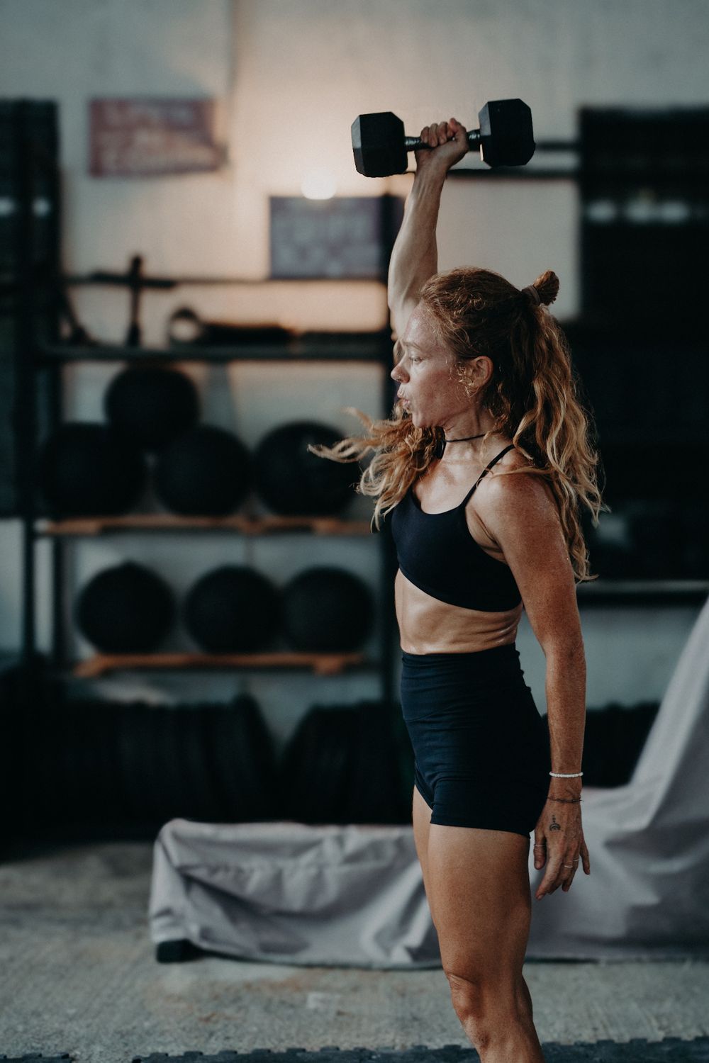 Fit woman in black sportswear lifts dumbbell overhead in a well-equipped gym.