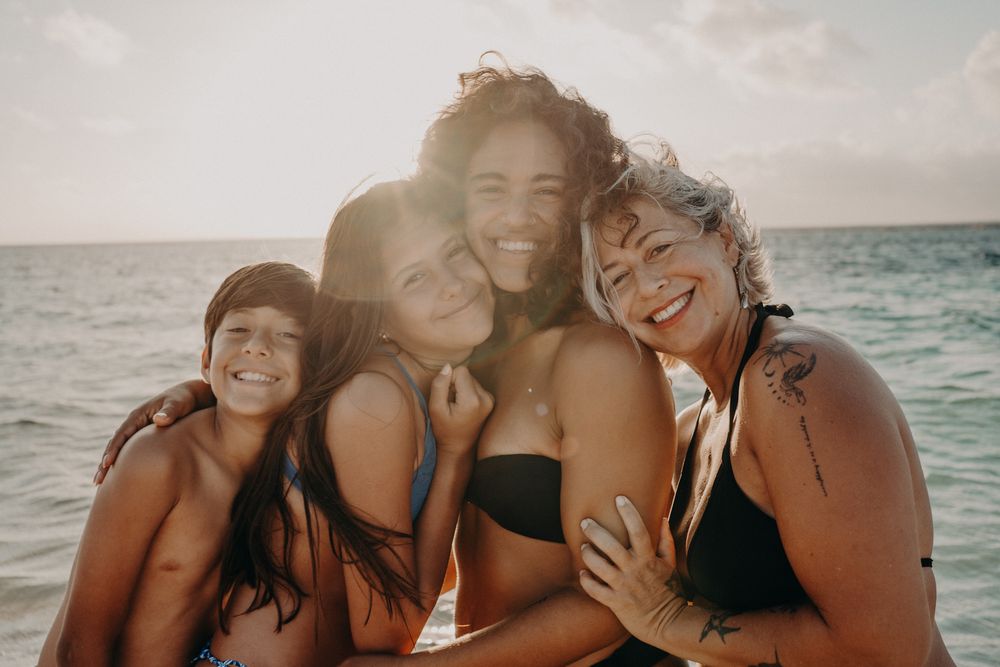 Four women smiling and hugging together on a beach at sunset with ocean in the background.