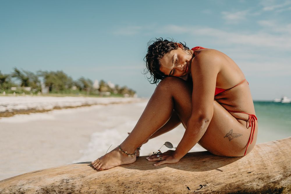 Woman in red swimwear sitting contentedly on a tropical beach with ocean and palm trees in background photoshoot in Puerto Morelos.