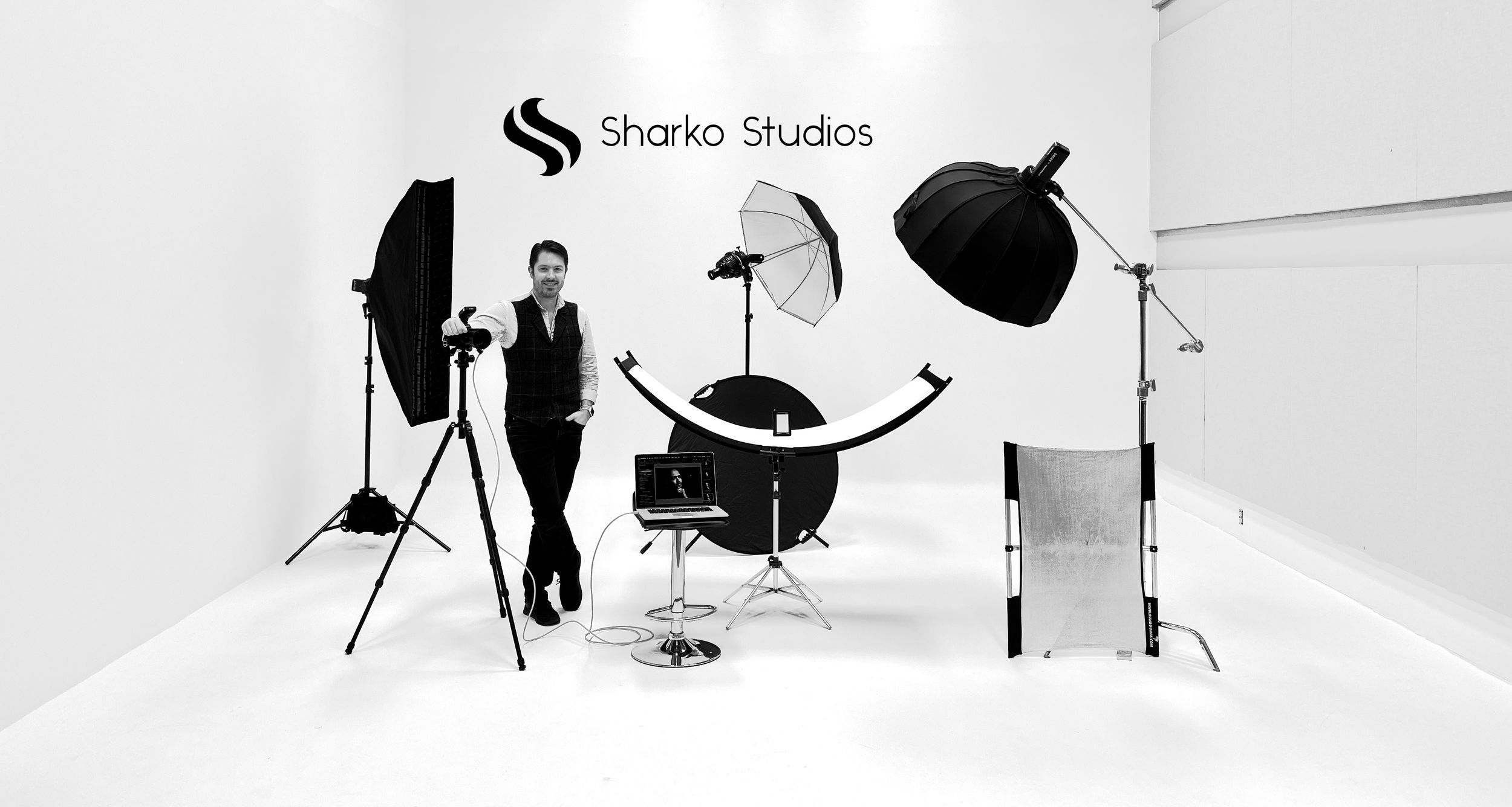 Evan Will of Sharko Studios in a black and white photo studio with lighting equipment, chair, and camera against a white backdrop.