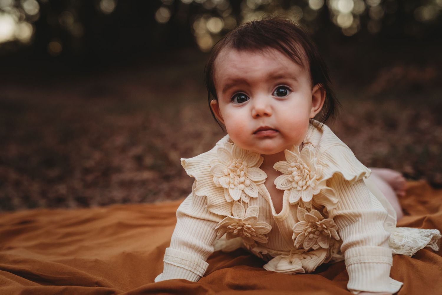 Small baby wearing cream colored ruffled outfit lays on brown blanket during outdoor autumn photoshoot.