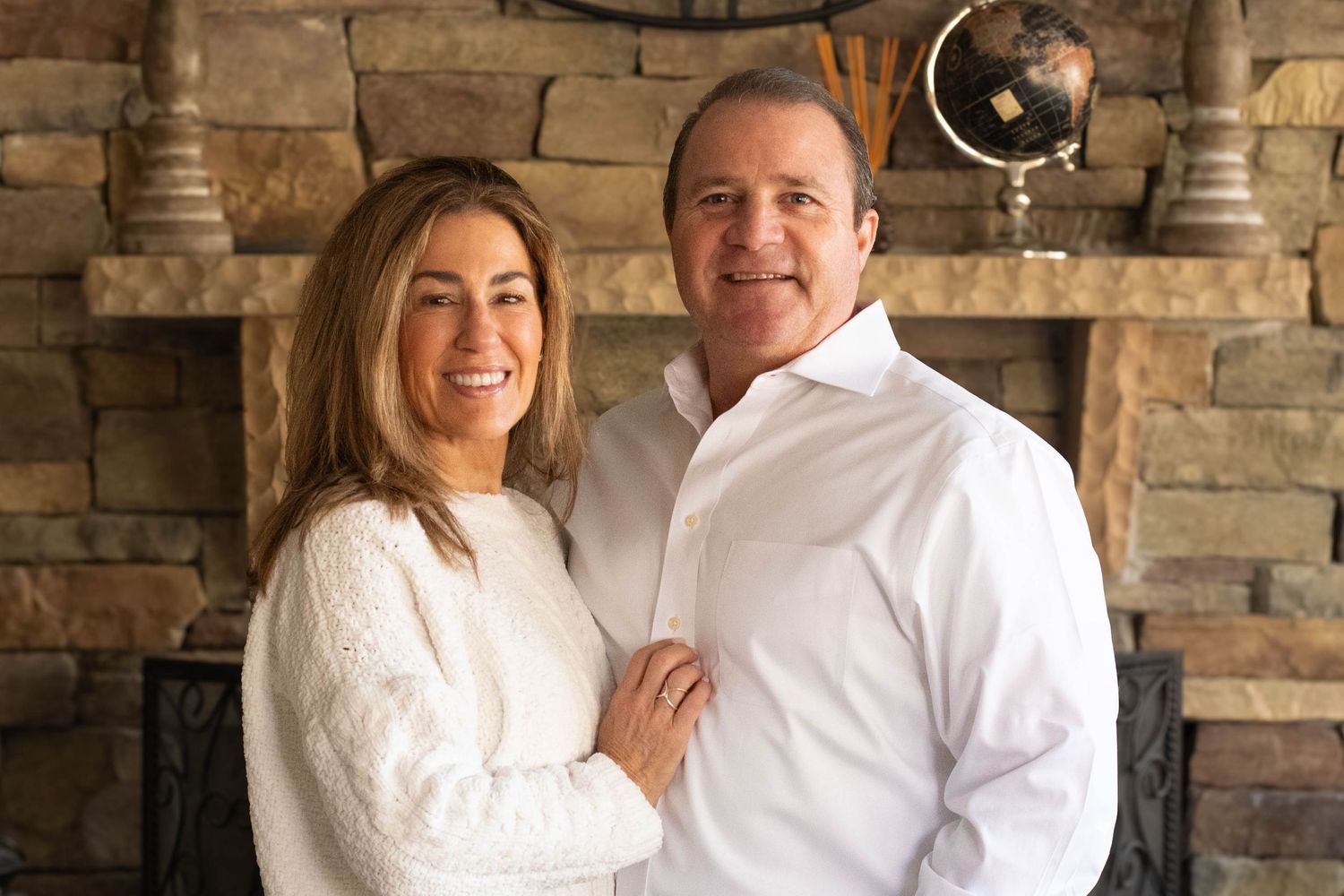 A couple poses together in front of a stone fireplace wearing white sweaters.