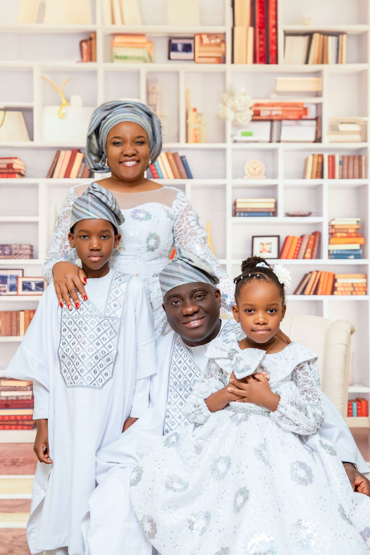 Family portrait in matching white traditional outfits against bookshelf background.