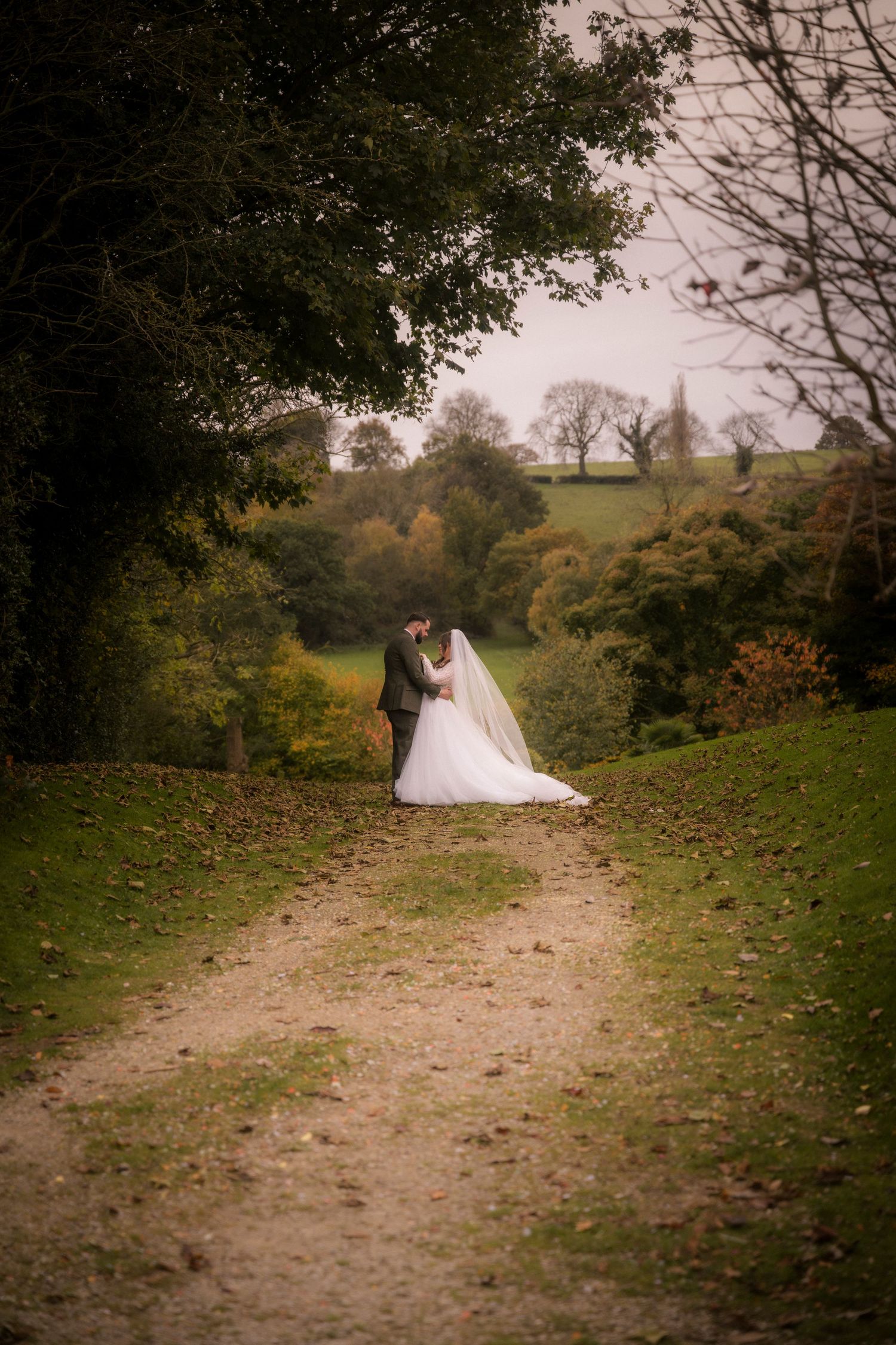A couple in wedding attire stand together on a rustic dirt path surrounded by autumn trees and rolling hills.