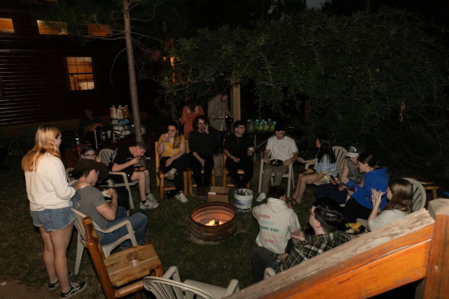 Group of people gathered around outdoor fire pit at night in backyard setting.