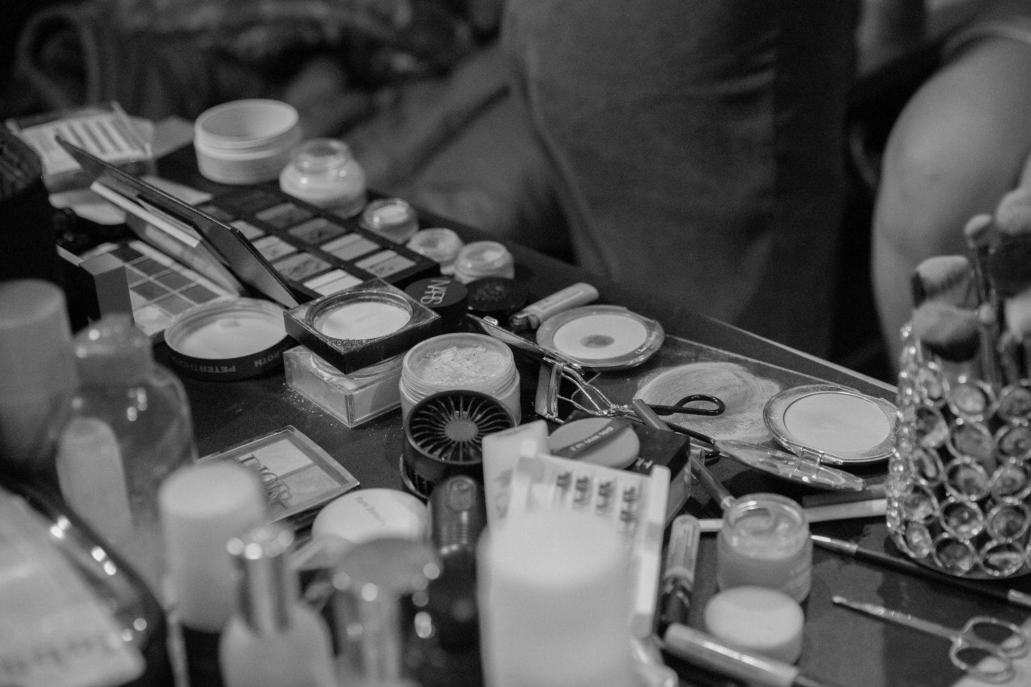 Black and white close-up of a bridal makeup station with cosmetics, brushes, and beauty tools arranged on a table.