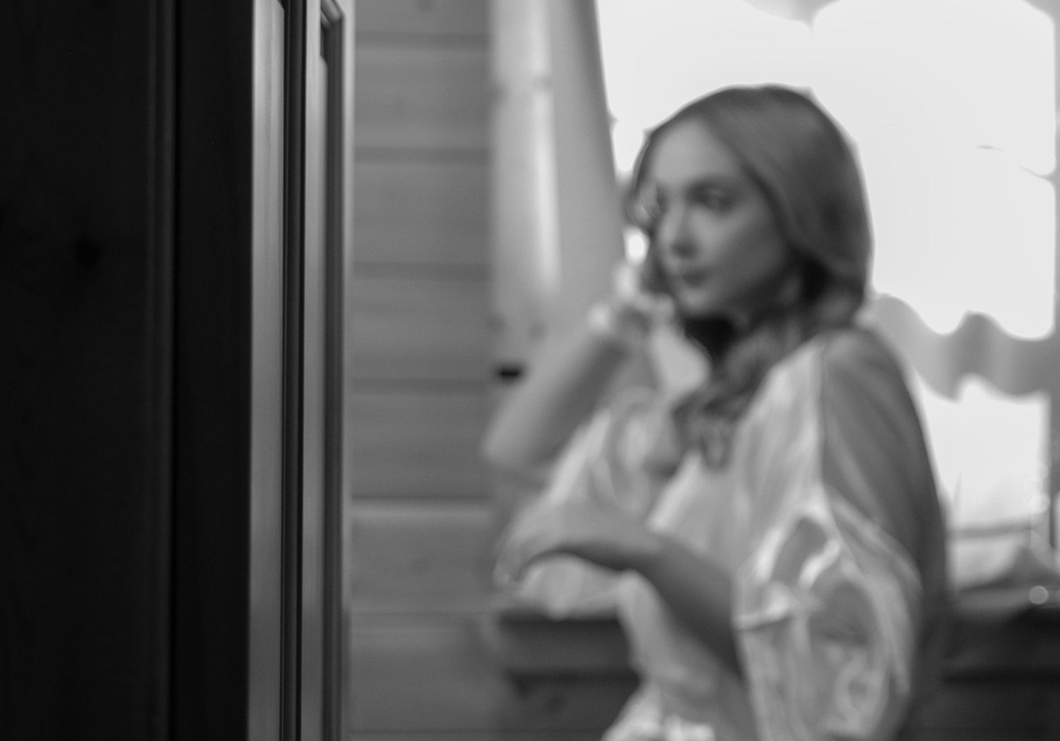 Black and white photo of a bride in a robe gazing out a window during wedding day preparations.
