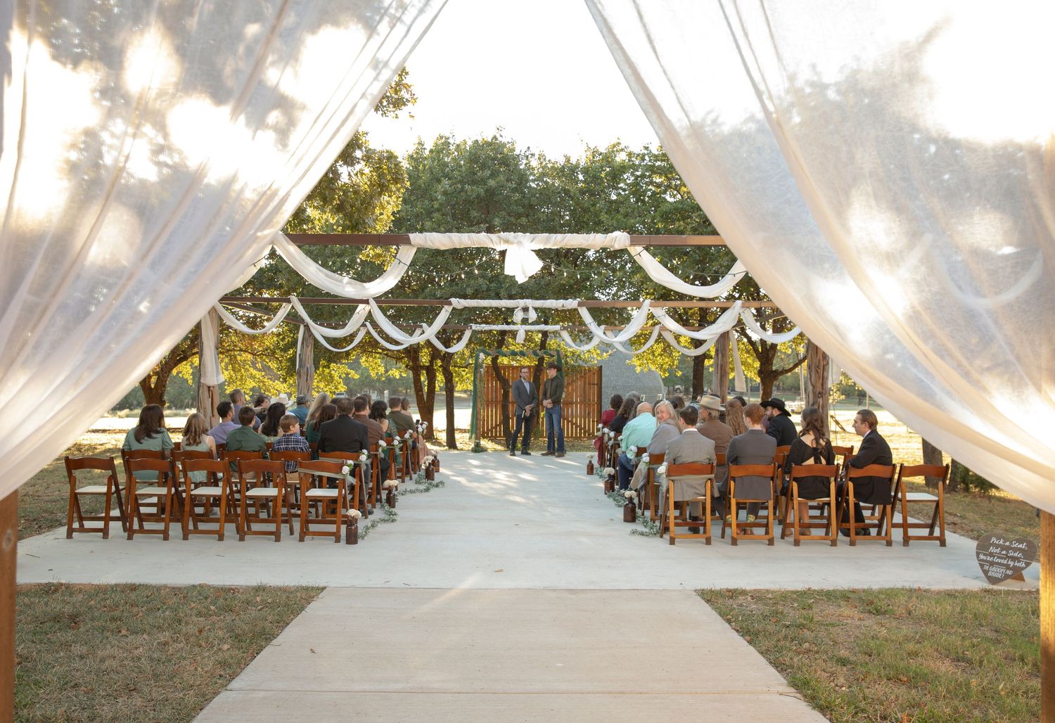 Elegant outdoor wedding ceremony with white draped arbor, wooden chairs, and guests seated along a white aisle.