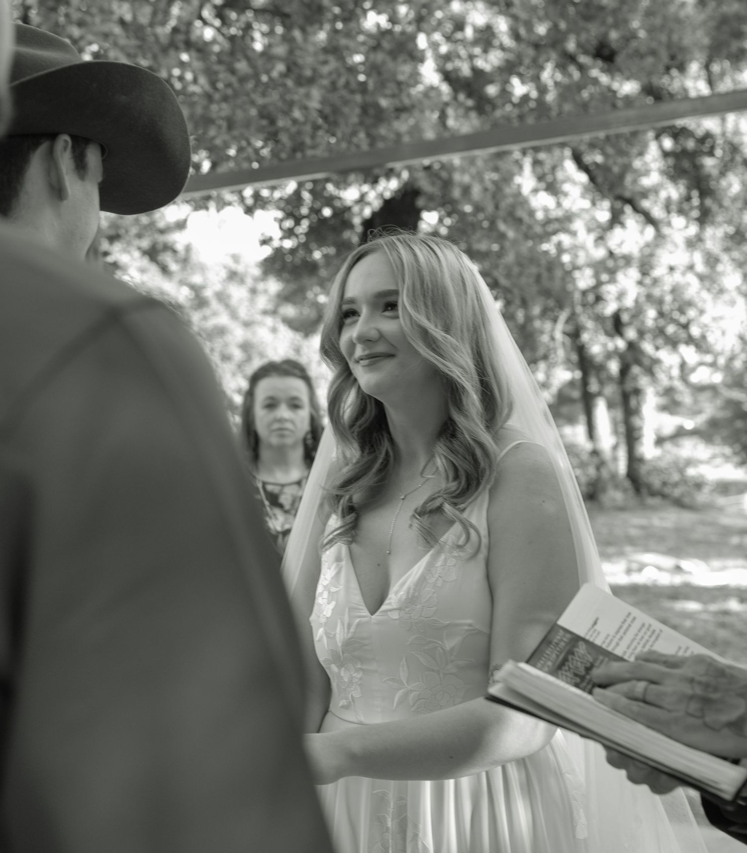 Black and white photo of an emotional bride smiling during her outdoor wedding ceremony beneath a veil.