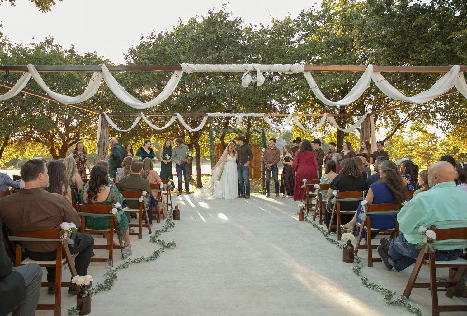 Outdoor rustic wedding ceremony with guests seated in wooden chairs under a white draped arbor among trees.