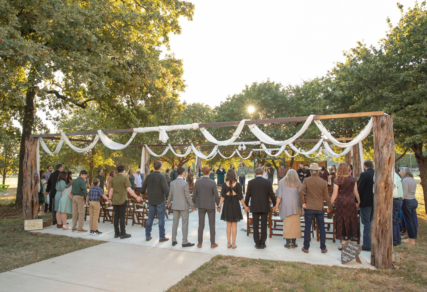 Outdoor rustic wedding ceremony under a wooden arbor draped with white fabric and lights surrounded by guests.