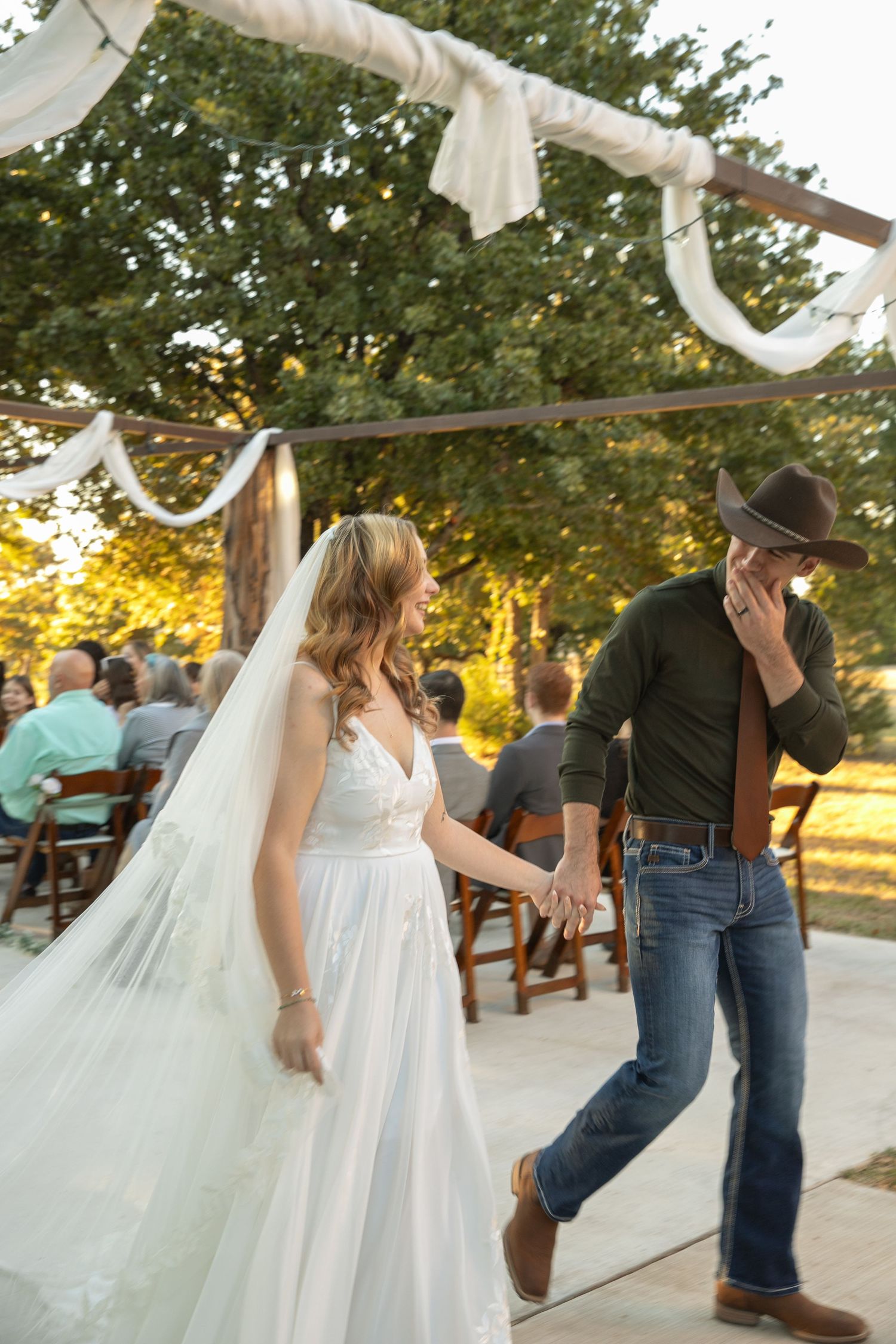 Bride in white gown and groom in cowboy hat walking after outdoor wedding ceremony.