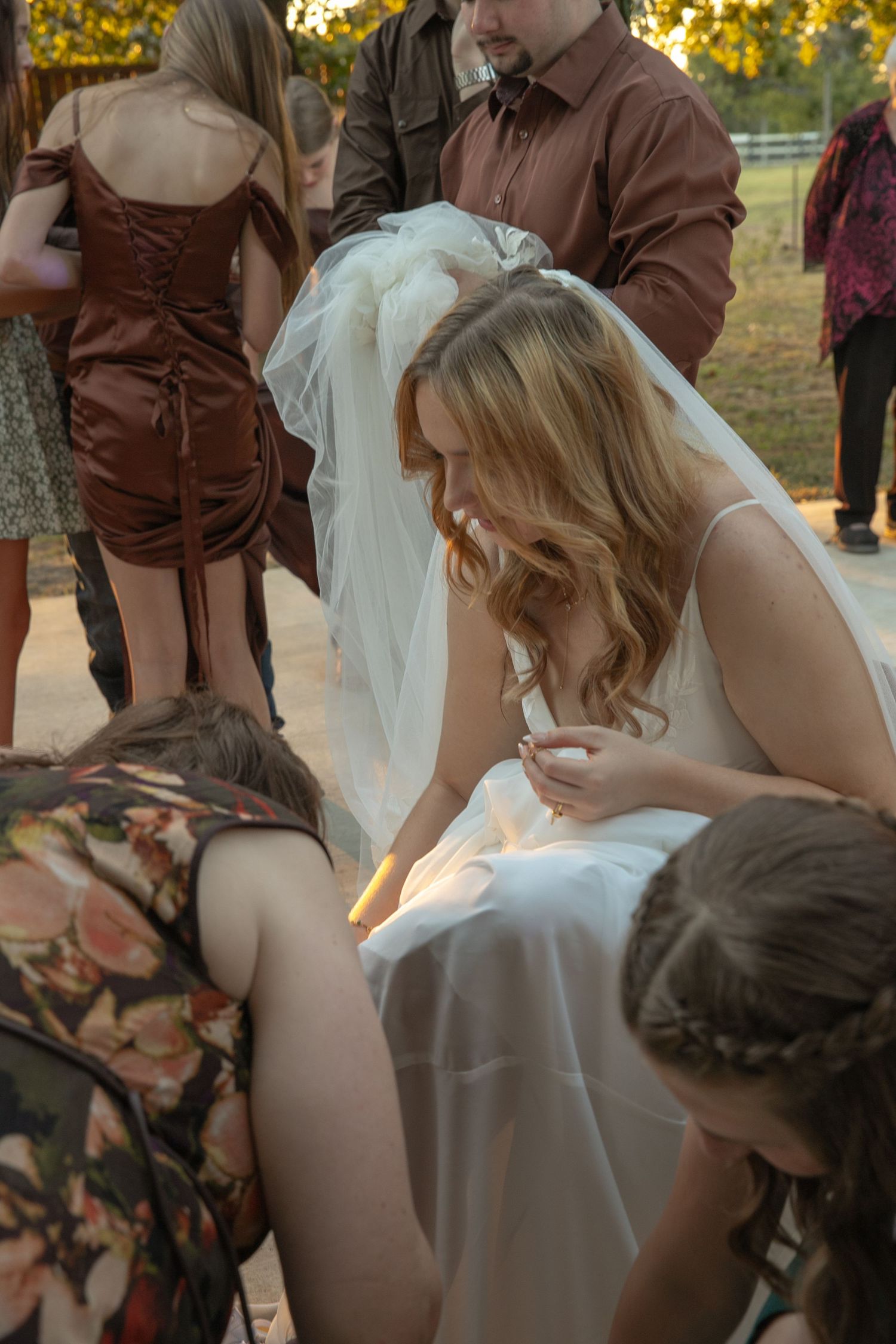 Bride in white wedding gown bending down with guests helping adjust her dress at an outdoor wedding reception.