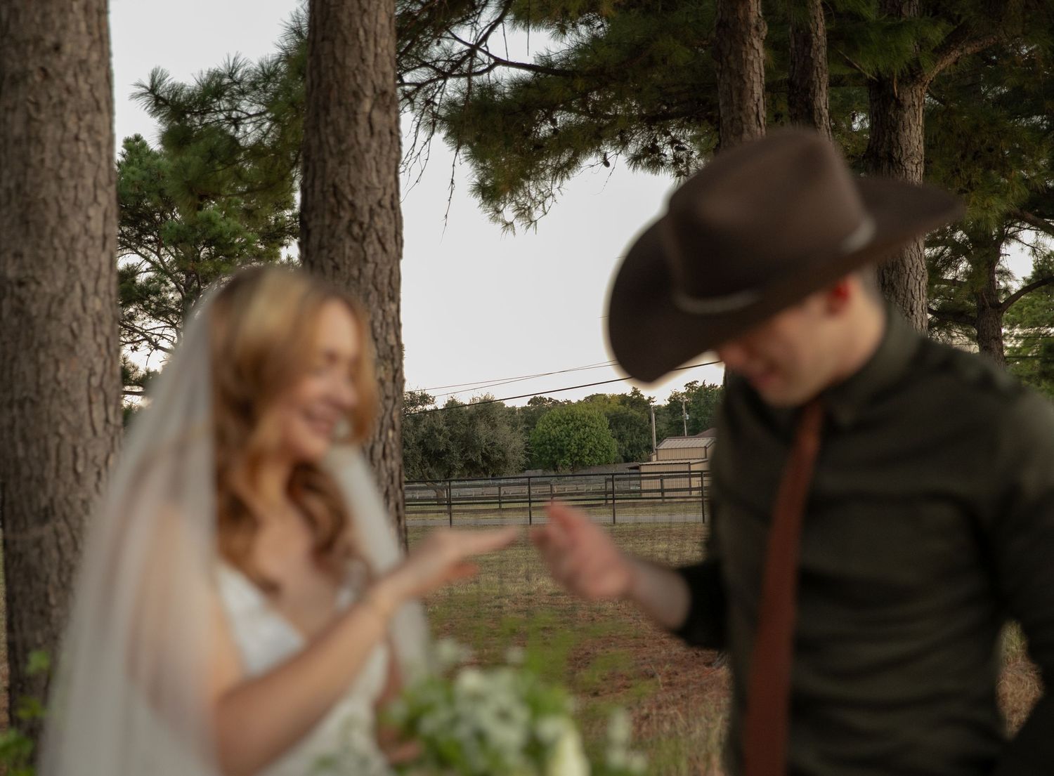 Bride smiling as groom in brown cowboy hat adjusts his hat near pine trees.