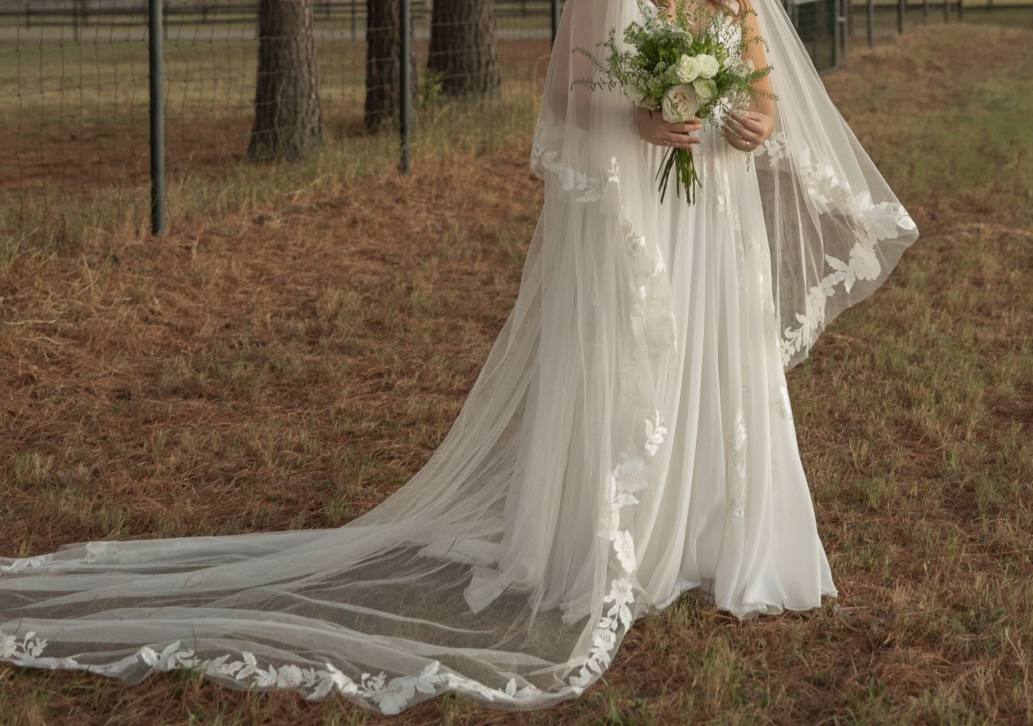 Elegant bride in a white lace-trimmed cathedral veil and gown holding a bouquet in a rustic outdoor field.